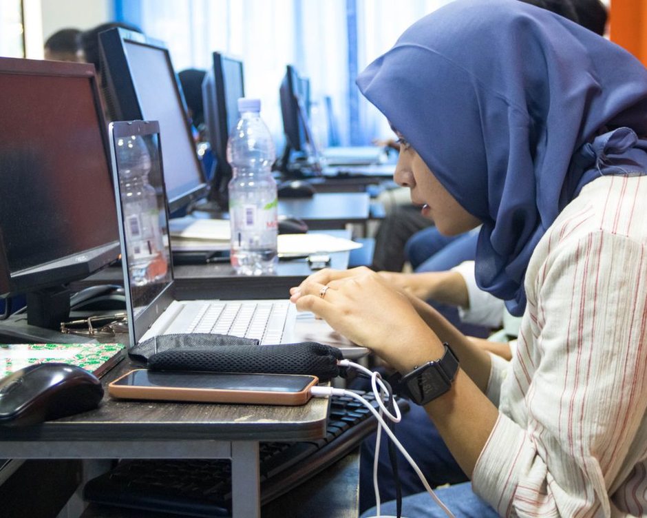 A student in a hijab coding on a laptop in a computer lab